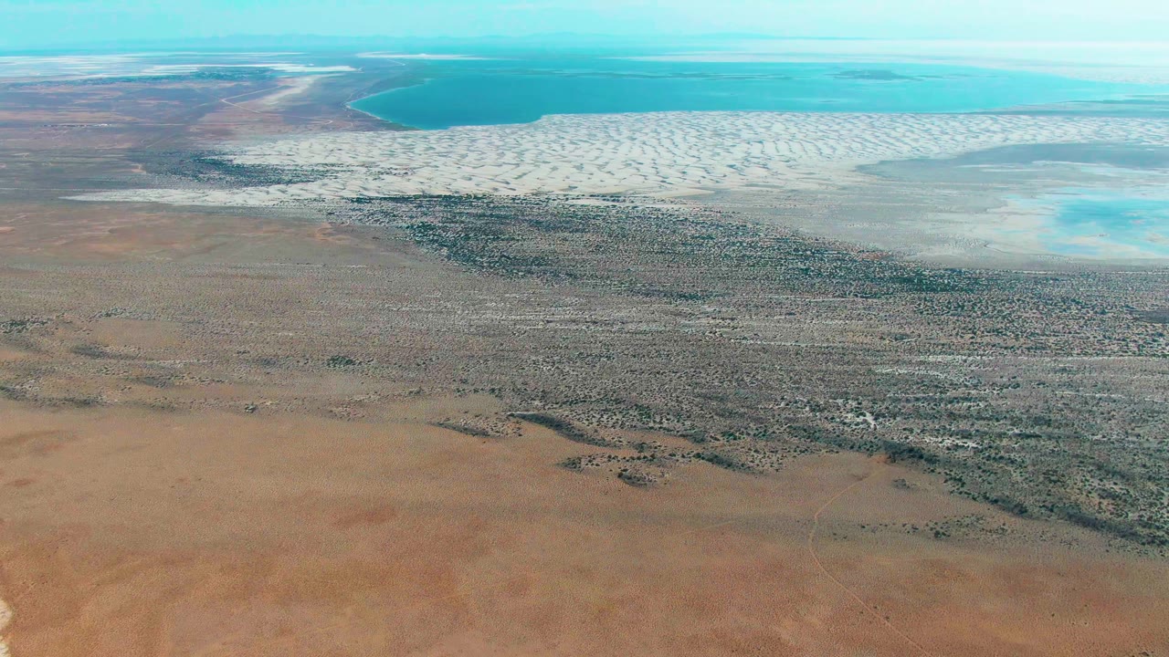 Aerial frame over Guerrero Negro showing brown desert foreground, pale patterned salt flats, turquoise shallow water near the horizon, blue sky, and no visible people.
