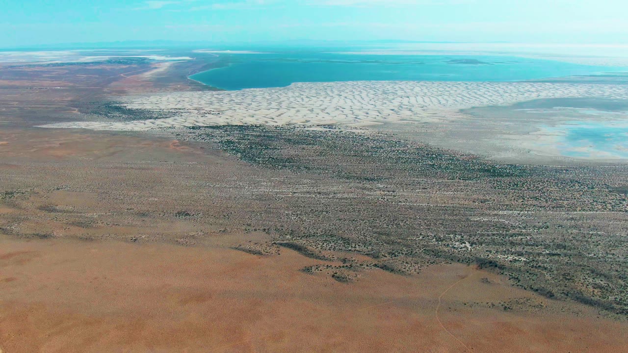 Wide aerial of arid terrain leading to patterned salt flats and shallow blue water.