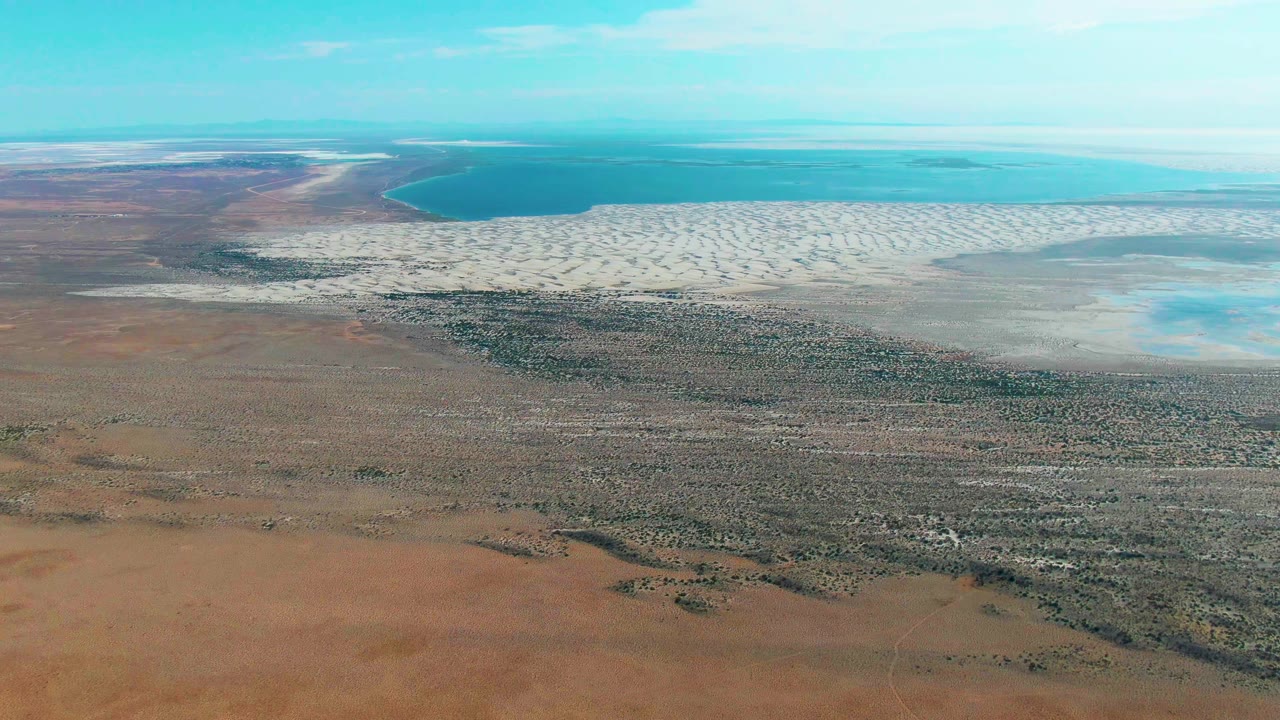 High aerial frame over Guerrero Negro with darker scrub-like patches across dry terrain, pale salt flats at midframe, blue-green water in the background, and no visible people.