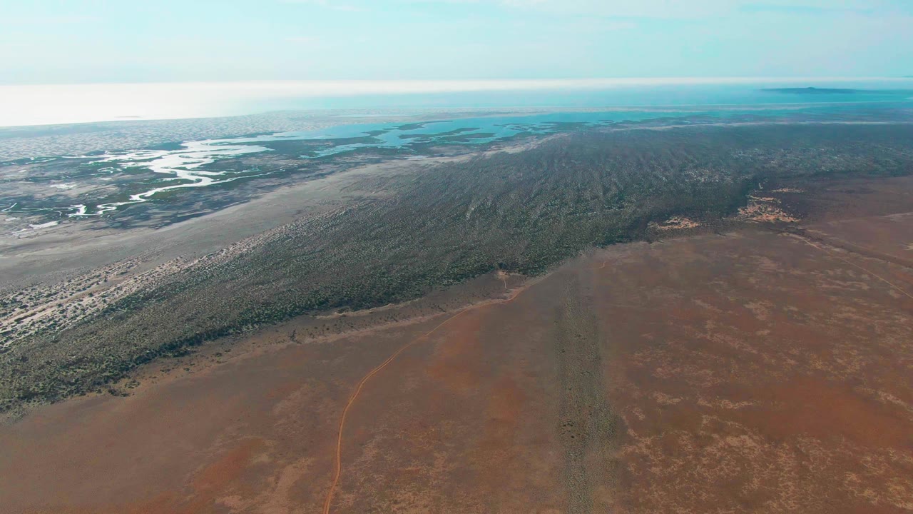 High aerial frame from Guerrero Negro, Baja California Sur, with red-brown ground across the lower frame, dense green vegetation running diagonally, light river channels to the left, blue coastal water behind, and no visible people.