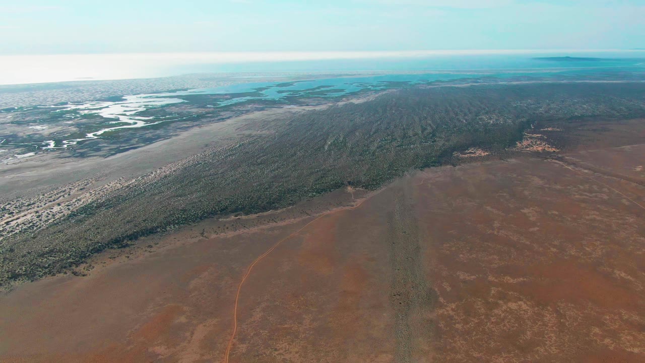 A later aerial frame in Guerrero Negro, Baja California Sur, showing the reddish plain filling more of the foreground, a dark forest edge across the center, bright meandering channels at upper left, and no visible people.
