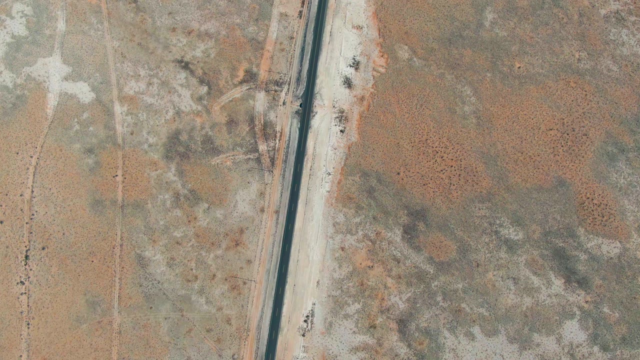 High overhead view of a straight road crossing dry brown and gray terrain in Guerrero Negro, Baja California Sur, with small vehicles on the pavement and no visible people.