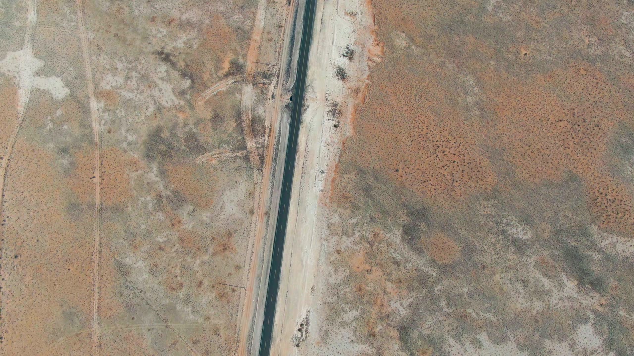 Aerial frame of the Guerrero Negro desert road with pale dirt shoulders, mottled brown ground, faint tracks, and no visible people.