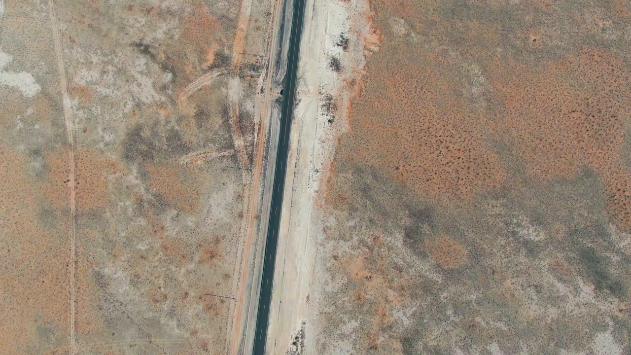 The road remains near the left side of the overhead frame while patchy arid land spreads across the right side in Guerrero Negro, with no visible people.