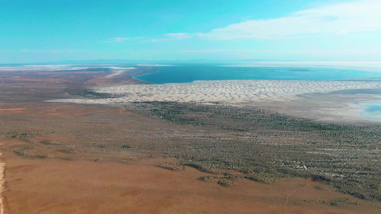 Wide aerial frame over Guerrero Negro, Baja California Sur, showing brown desert scrub in front, pale tidal flats at the shore, blue ocean beyond, and no visible people.