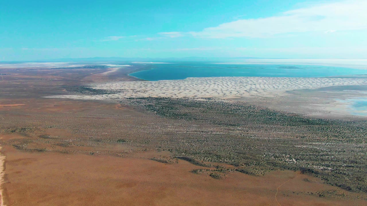 Aerial view in Guerrero Negro with reddish-brown land in the foreground, green scrub bands through the middle, a pale coastal strip, blue ocean, and no visible people.