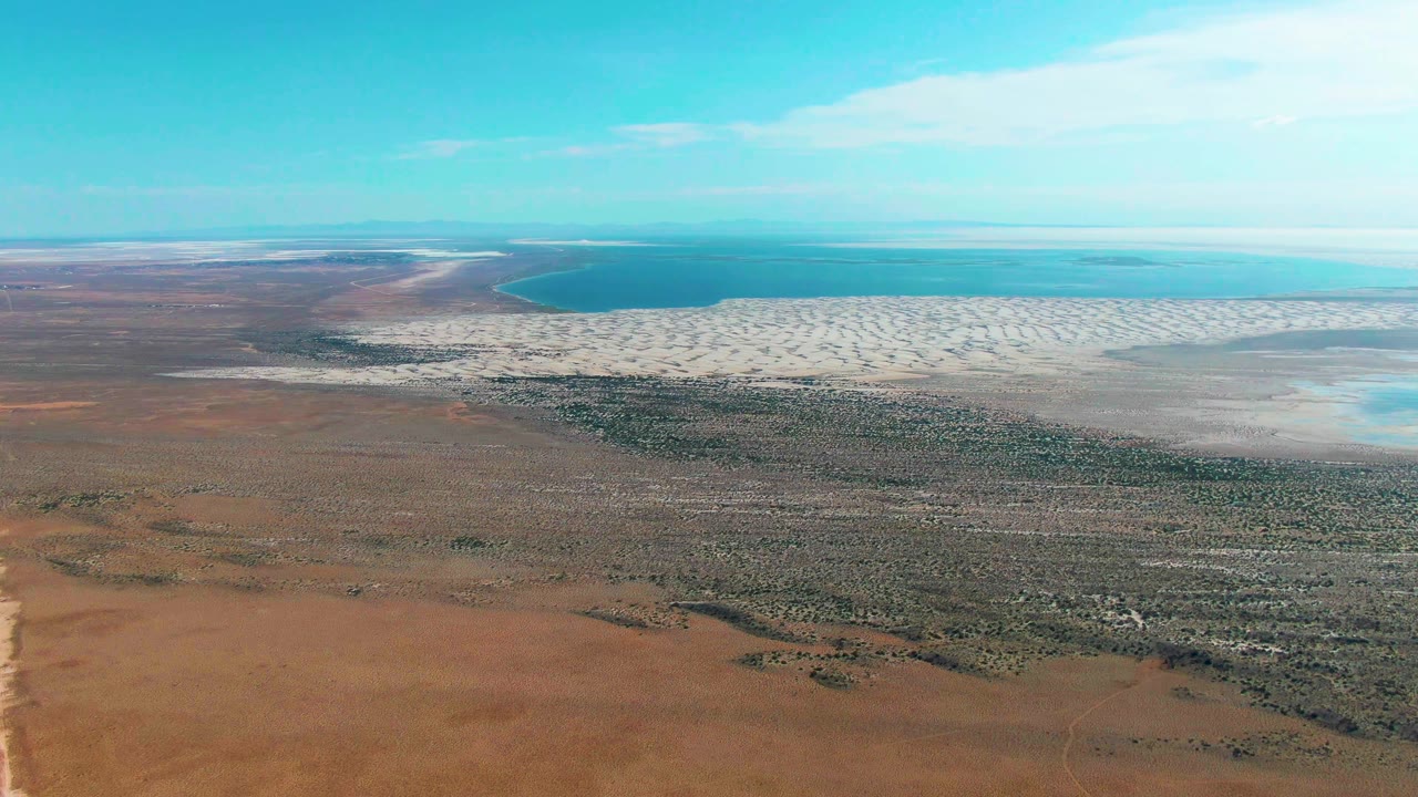 High drone-style frame over Guerrero Negro, Baja California Sur, with desert scrub below, white shoreline textures across the midground, blue ocean on the horizon, and no visible people.