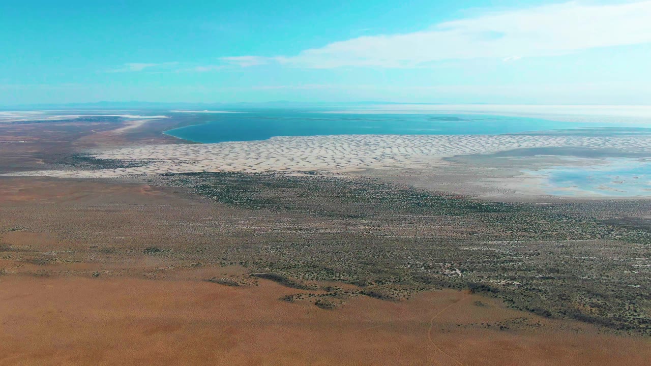 Opening aerial frame over Guerrero Negro, Baja California Sur, Mexico, with red desert ground below, sparse green vegetation, pale patterned shallows, blue water near the horizon, and no visible people.