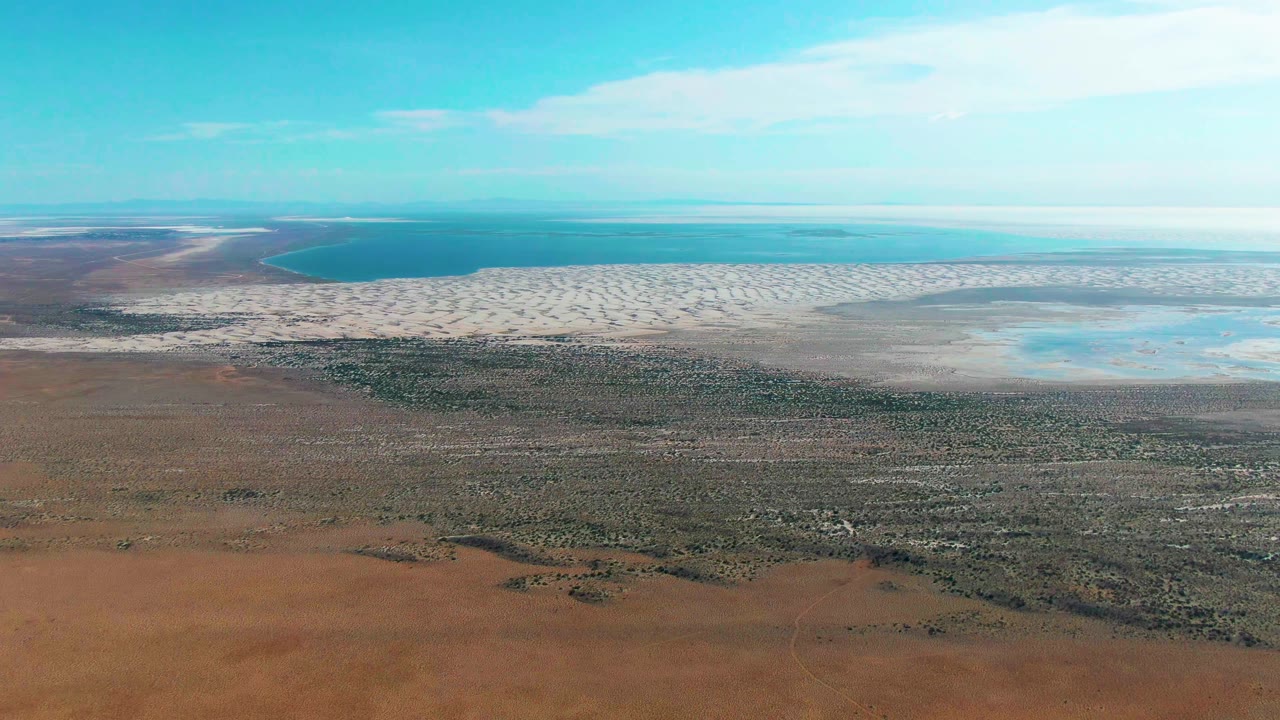 Wide aerial frame over Guerrero Negro showing reddish earth in the lower frame, scrubby green vegetation across the middle, white patterned flats and shallow water behind it, and no visible people.