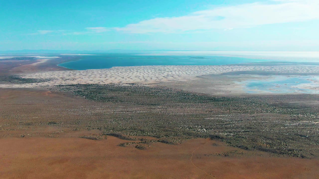 Aerial shoreline view with dry terrain, sparse vegetation, pale shallows, and turquoise water.