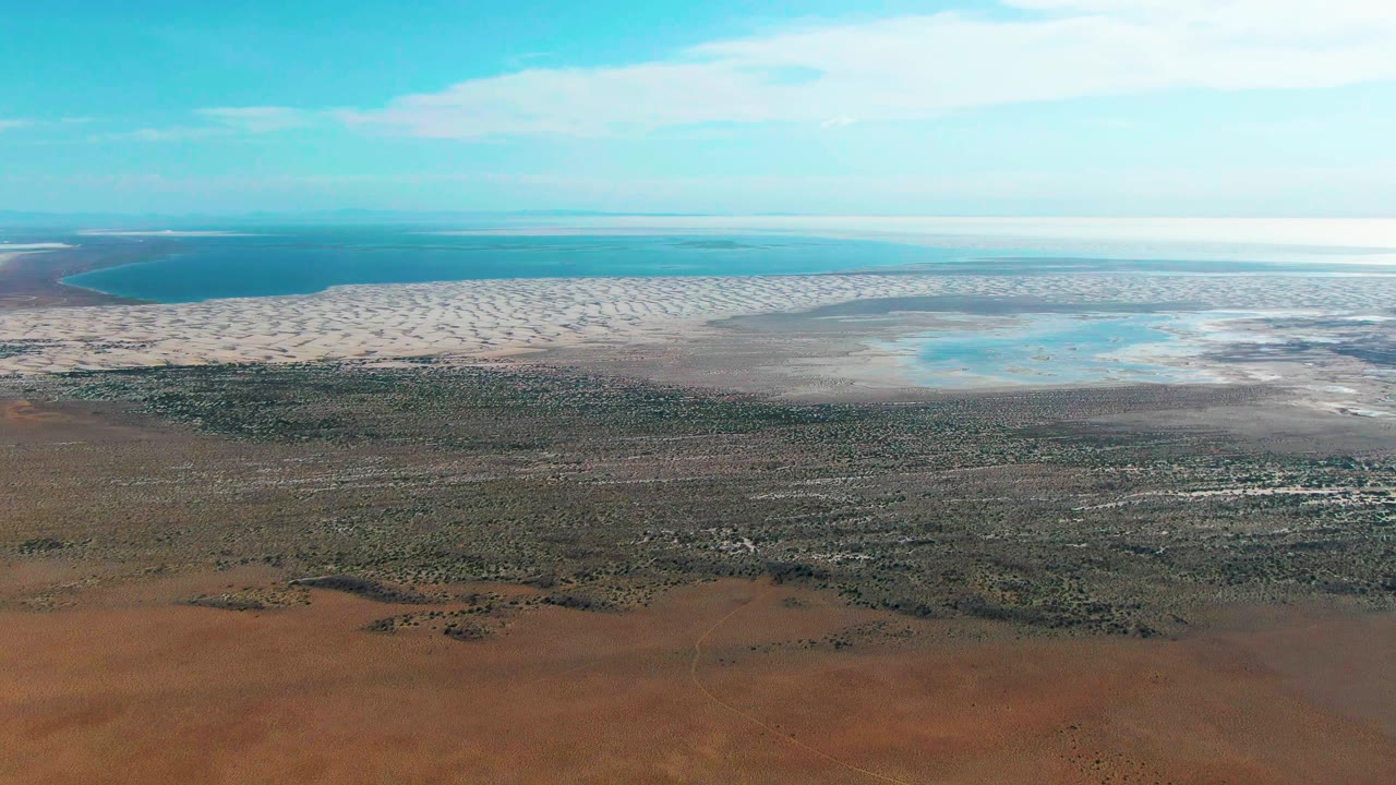 Aerial view of red earth, sparse vegetation, tidal flats, ocean, and blue sky.