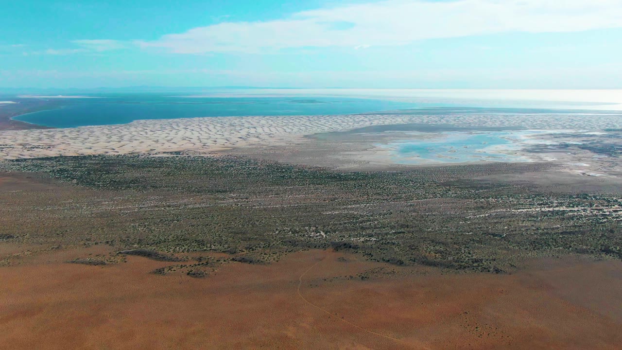 Aerial view in Guerrero Negro, Baja California Sur showing more of the pale tidal-flat band across the middle, reddish ground below, distant water, blue sky, and no visible people.