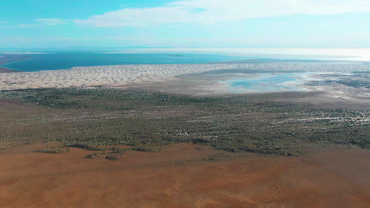 Wide coastal aerial over Guerrero Negro, Baja California Sur with patterned pale flats stretching across the upper frame, darker wet shoreline, red terrain below, and no visible people.