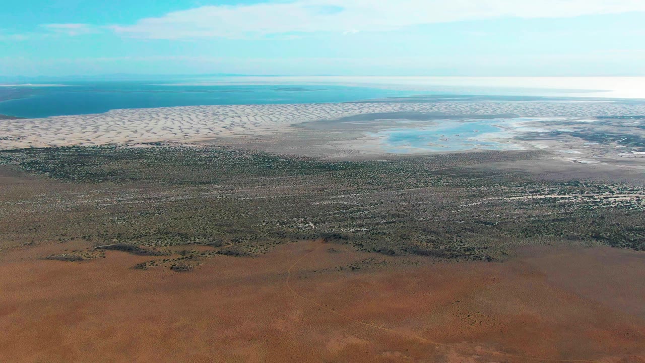 Guerrero Negro aerial frame with the horizon high over blue water, a broad pale tidal-flat strip, darker muddy shore, scrubby vegetation, red foreground, and no visible people.