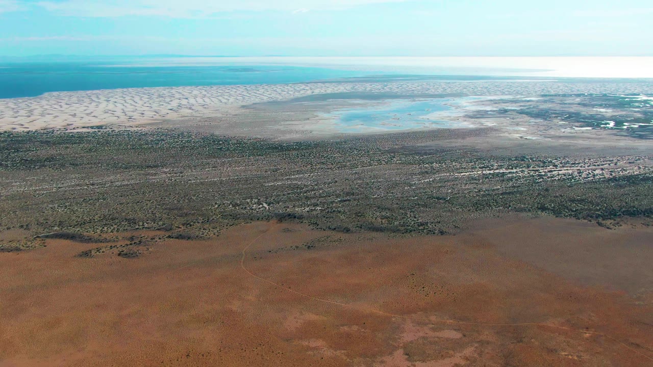 Aerial view of red-brown desert, green vegetation, pale dunes, and blue coastal water.