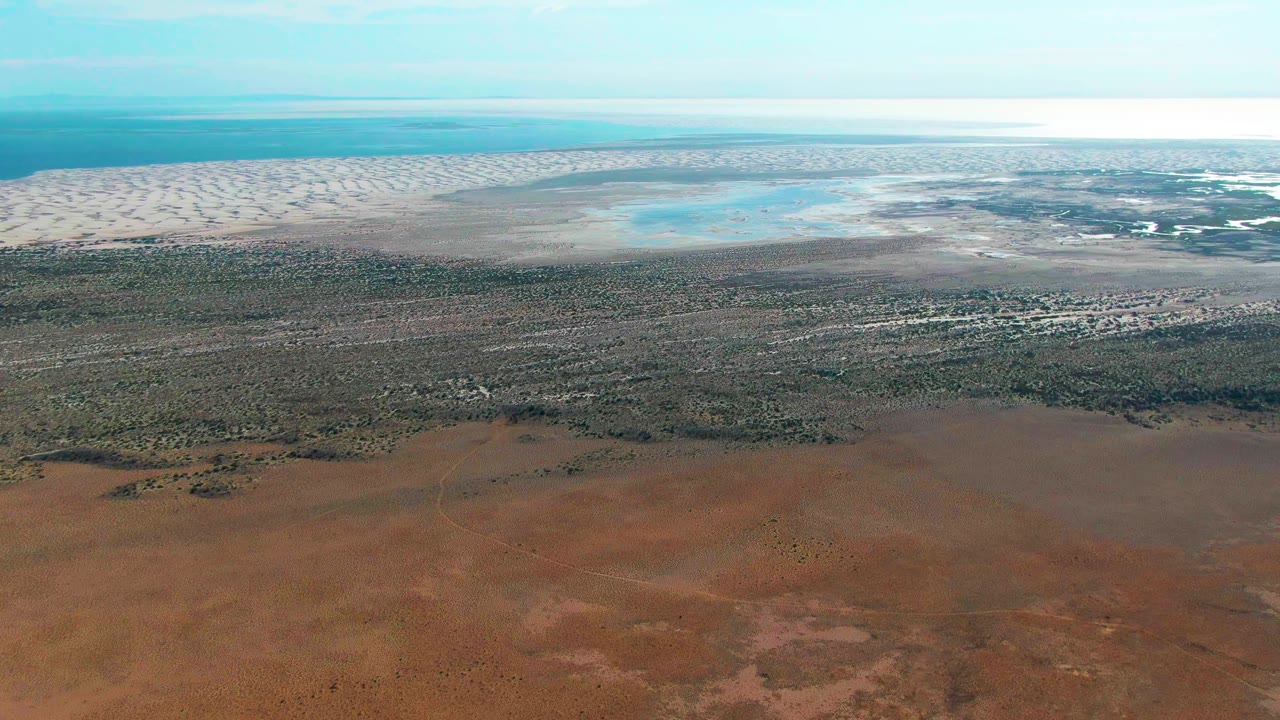 At 0:01, the Guerrero Negro aerial holds a broad view of reddish terrain in front, darker vegetation across the middle, pale sand flats, blue water, haze, and no visible people.