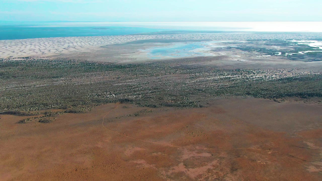 At 0:02, the aerial view over Guerrero Negro shows a wider sweep of scrubland and reddish soil below pale dunes and blue coastal water, with no visible people.