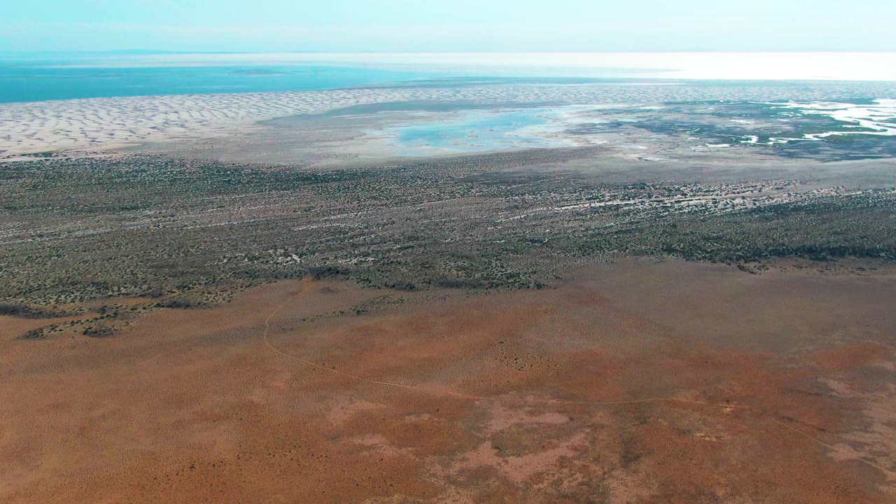 At 0:03, Guerrero Negro's coastline appears with red-brown foreground terrain, a long green band, pale dunes, blue water pools, bright haze, and no visible people.