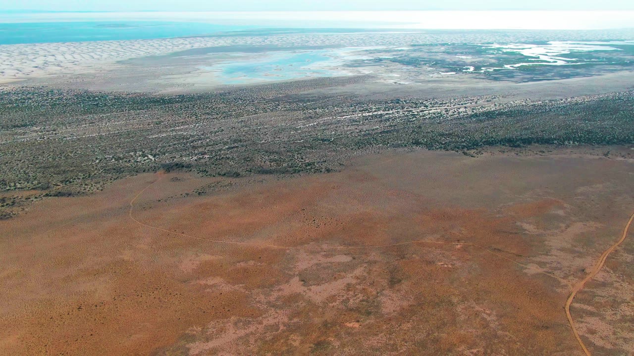 Aerial frame over Guerrero Negro, Baja California Sur, showing reddish dry terrain below, a vegetation band across the middle, pale water near the horizon, and no visible people.