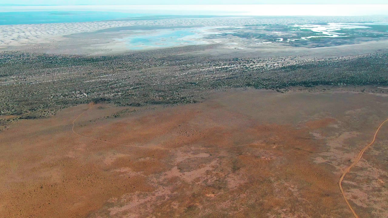 Aerial view in Guerrero Negro with dry reddish foreground, mottled scrub and vegetation in the midground, bright water and pale flats beyond, and no visible people.