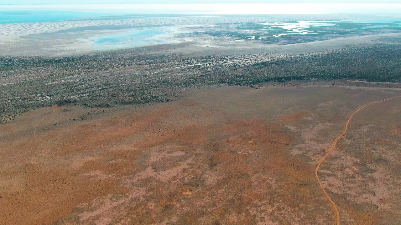 A high aerial frame over Guerrero Negro, Baja California Sur shows a narrow dirt road through reddish brown arid ground, darker vegetation in the distance, pale water near the horizon, and no visible people.