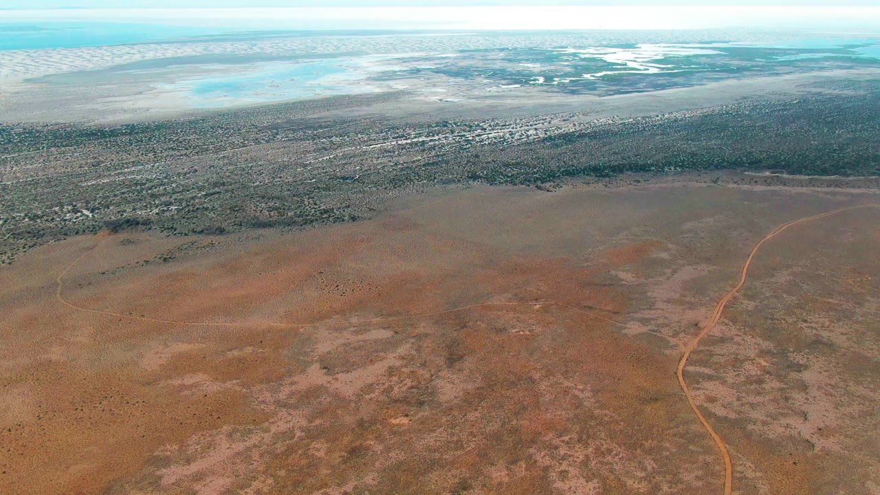 At 0:01, the aerial view over Guerrero Negro, Baja California Sur keeps the dirt road near the right side of the frame, with mottled dry earth below, a dark scrub line beyond it, pale water, and no visible people.