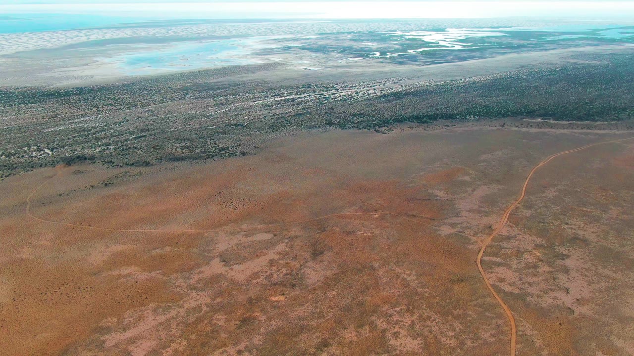 At 0:02, the Guerrero Negro aerial frame shows the dirt track running up the right half of the dry terrain, with textured brown ground, a darker vegetation strip, light blue water at the top, and no visible people.