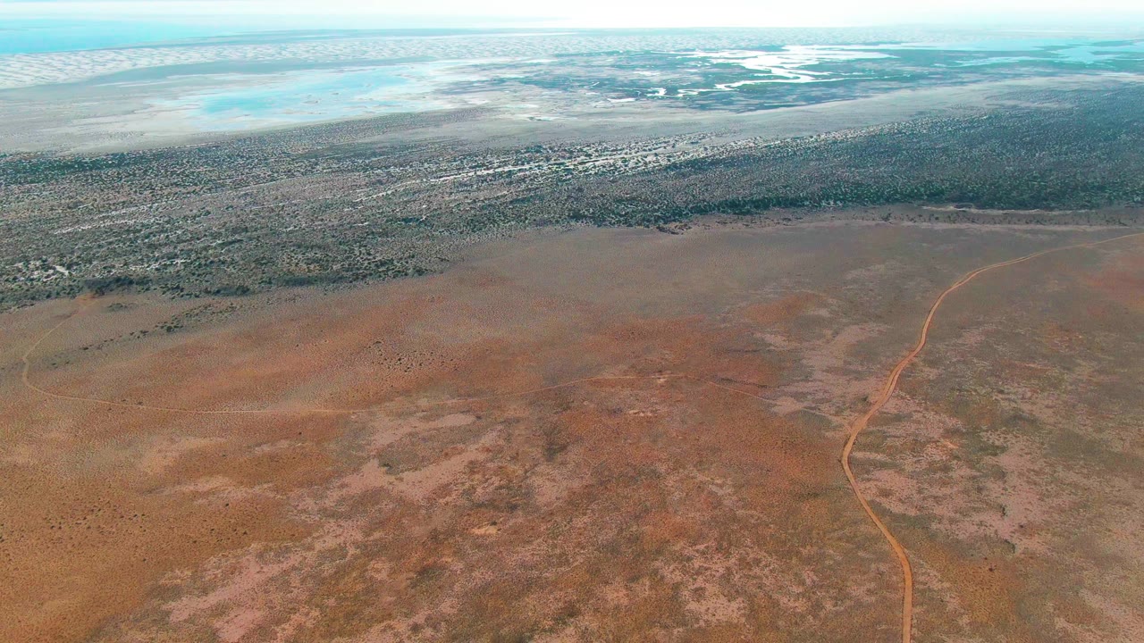 Aerial landscape with a dirt road crossing dry terrain below distant scrub and water.