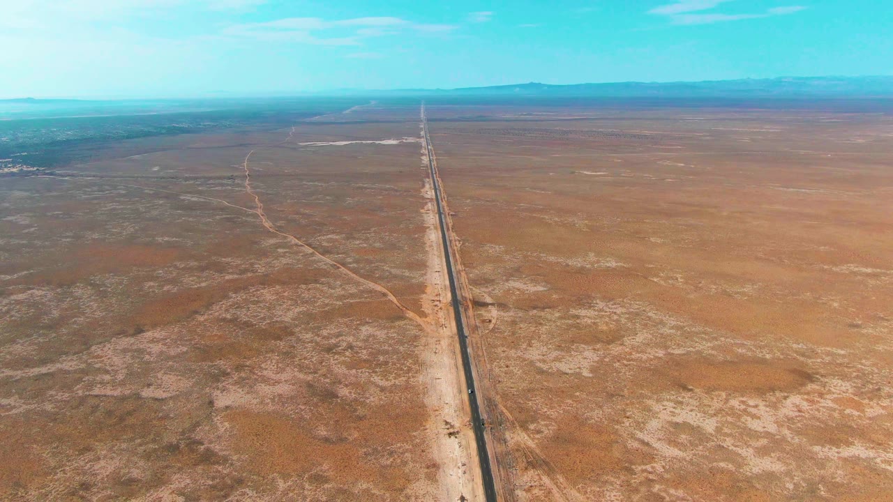 Mid-clip aerial frame over Guerrero Negro, Baja California Sur, showing the road cutting upward through sandy desert patches beneath a clear blue sky, with no visible people.