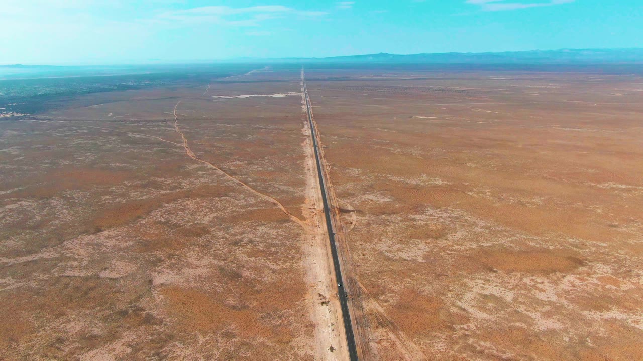 Later aerial frame in Guerrero Negro, Baja California Sur, with the paved road near center, a faint dirt track to one side, and low mountains across the horizon, with no visible people.