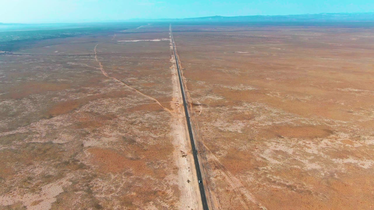 Aerial view in Guerrero Negro with the highway centered in the frame, pale roadside dirt on both sides, scattered desert scrub, distant low hills, and no visible people.