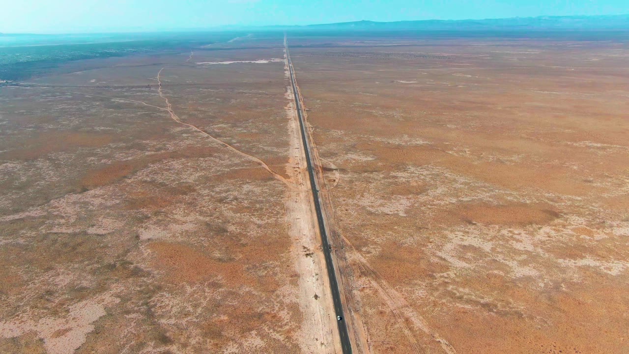 Aerial frame over Guerrero Negro where a dark road divides flat orange and tan desert, with faint dirt paths branching away and no visible people.