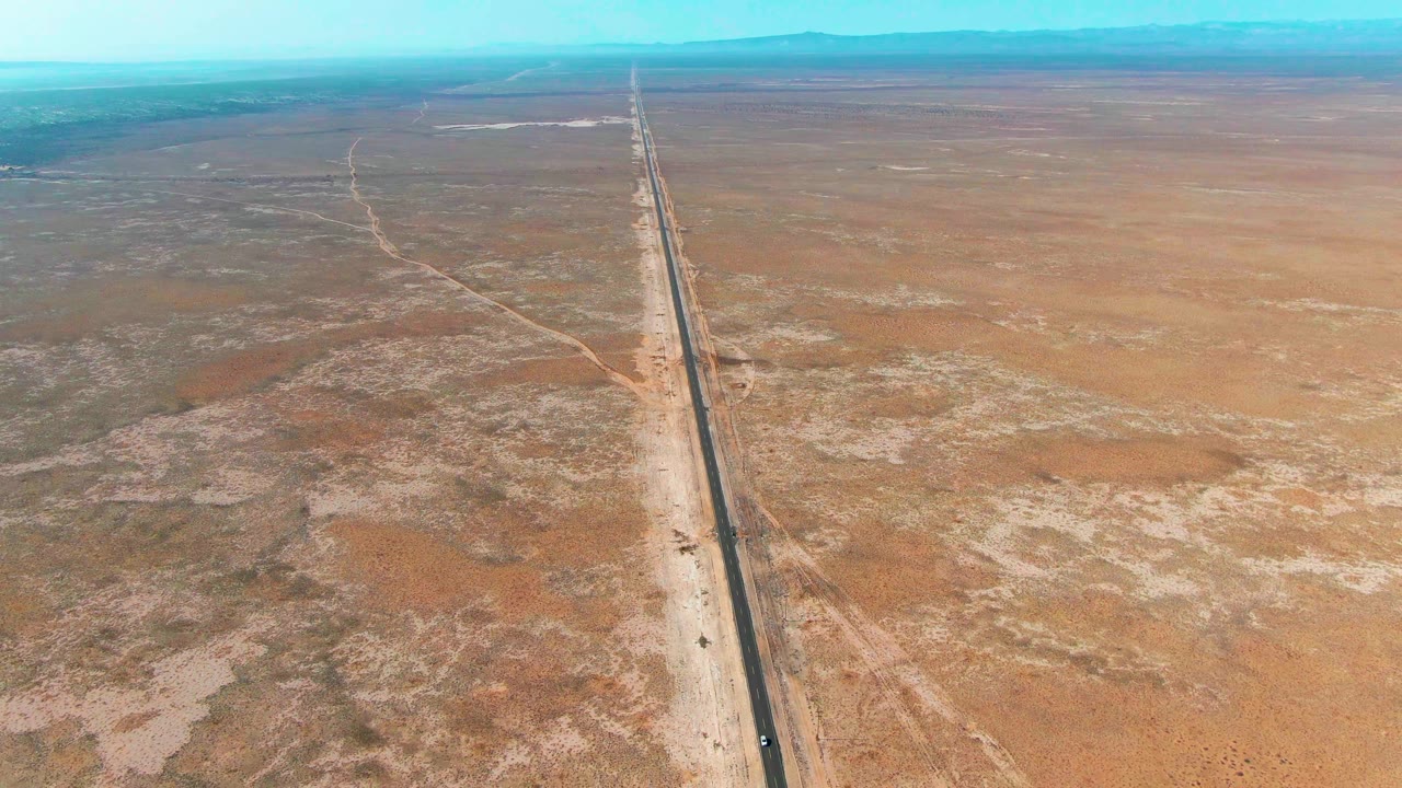 A high aerial view in Guerrero Negro shows the straight highway slightly right of center, broad scrubland to the left, open sandy ground to the right, and no visible people.