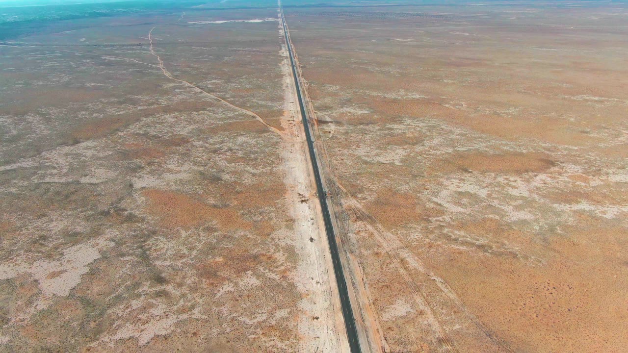 Drone-like aerial frame near Guerrero Negro showing the road running toward the horizon through mottled brown desert, with a small vehicle on the pavement and no visible people.