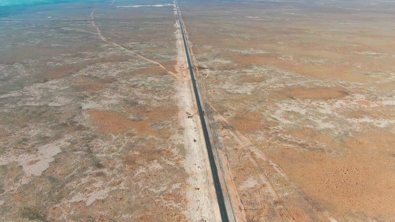High oblique view near Guerrero Negro with the straight road crossing the right side of frame, a small vehicle visible on the road, and broad brown desert with no visible people.