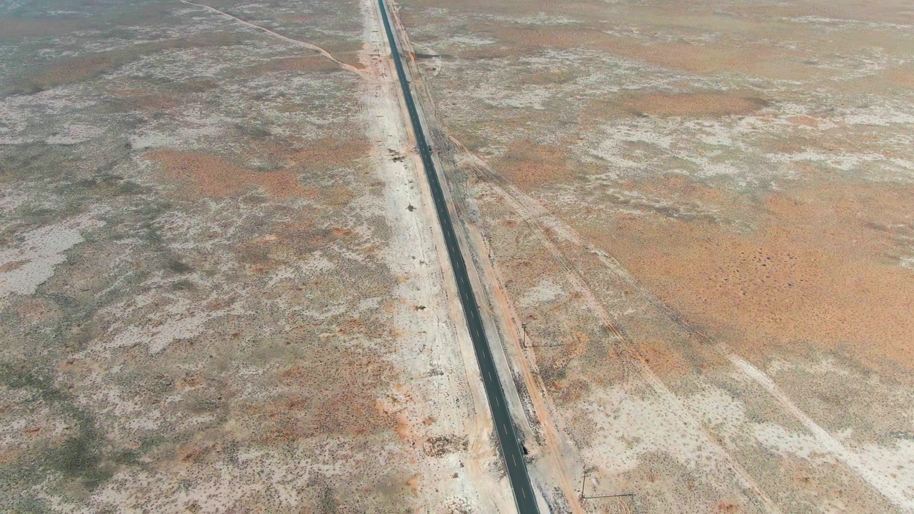 Aerial frame from Guerrero Negro, Baja California Sur, Mexico, showing a straight paved highway cutting through pale sandy desert with sparse vegetation and no visible people.