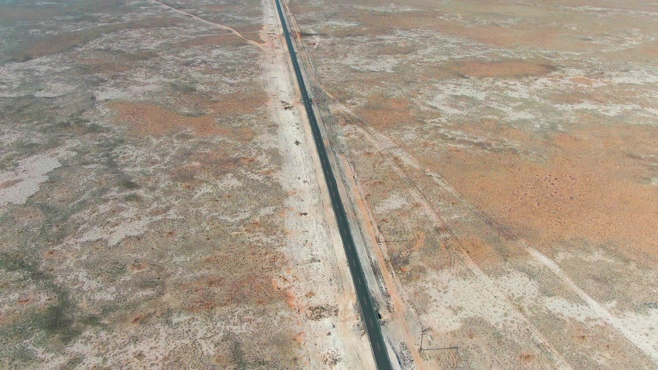 A second aerial frame in Guerrero Negro shows the highway slightly farther through mottled dry earth, orange sand patches, low scrub, and no visible people.