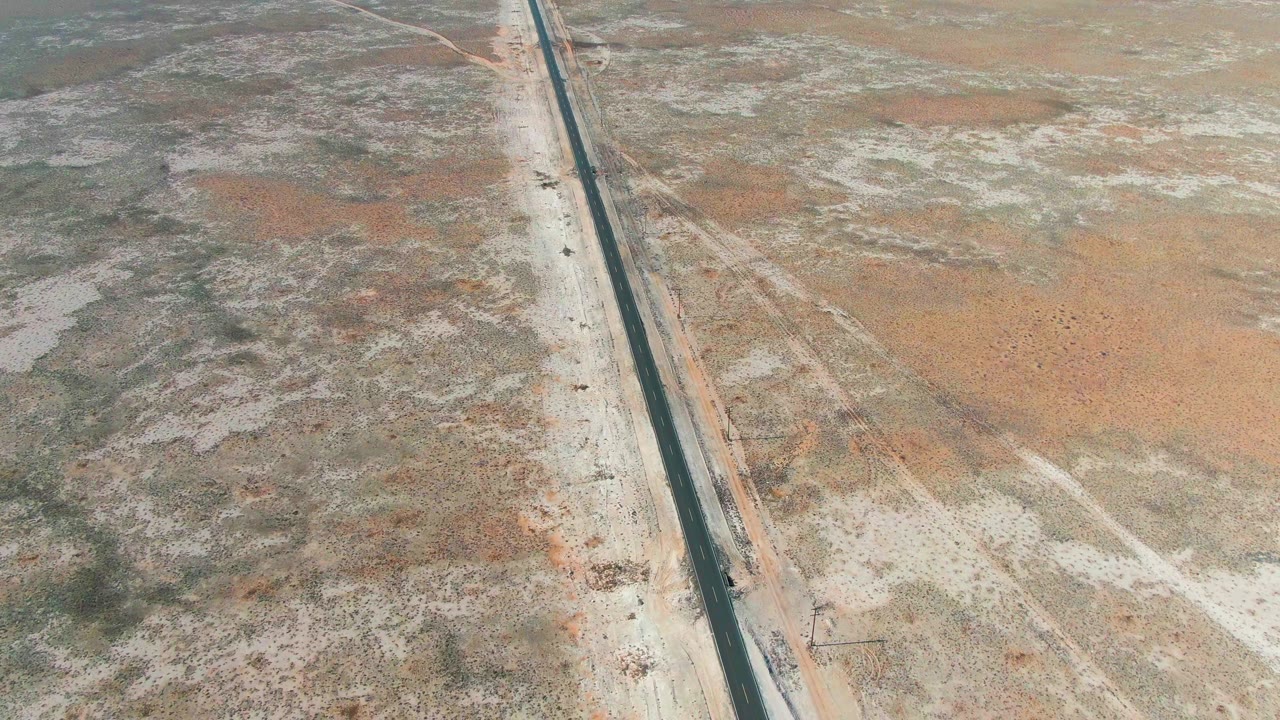 At 0:03, the aerial view in Guerrero Negro shows the vehicle lower along the straight road, with dry gray-green scrubland on one side, orange sand on the other, and no visible people.