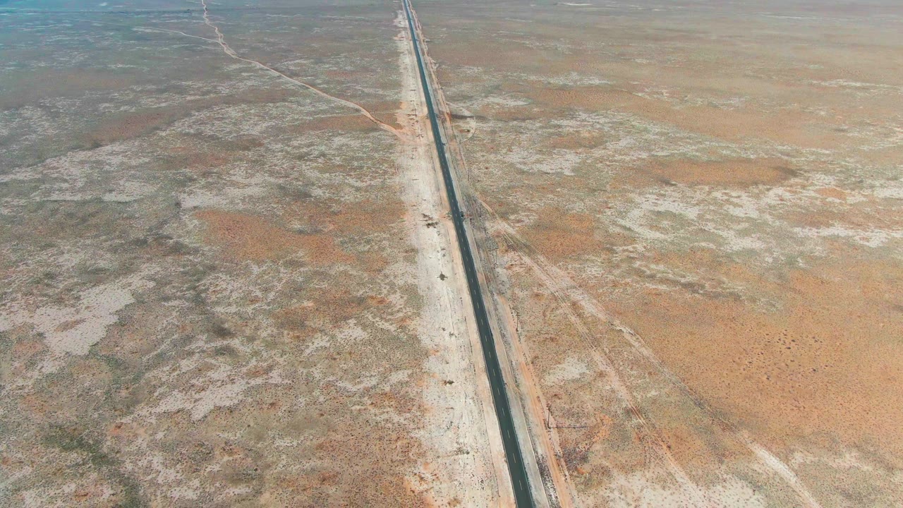 Overhead view in Guerrero Negro showing the road slightly right of center, bordered by pale shoulders, mottled scrub, reddish soil, and no visible people.