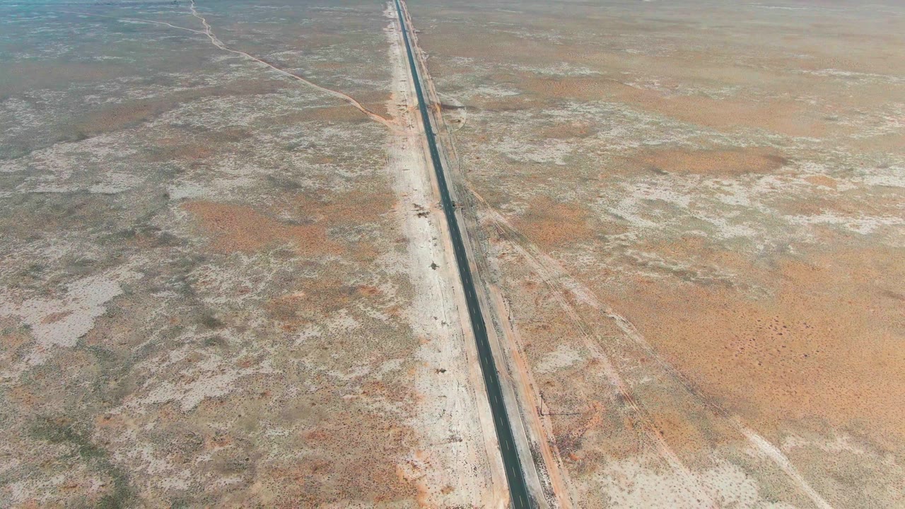Aerial view of the Guerrero Negro desert road running toward the distance through pale sandy terrain, rust-colored patches, sparse vegetation, and no visible people.