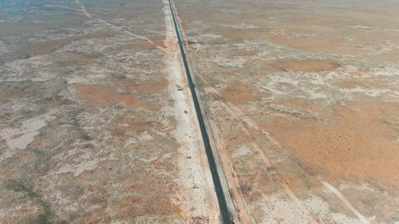 Drone-height view over Guerrero Negro with a small vehicle on the straight road, dry pale shoulders, reddish desert patches, faint side tracks, and no visible people.