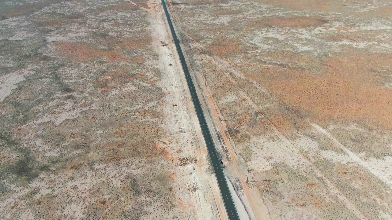 Aerial frame from Guerrero Negro, Baja California Sur, Mexico showing a straight dark highway crossing pale sandy desert with a small truck near the lower road section and no visible people.