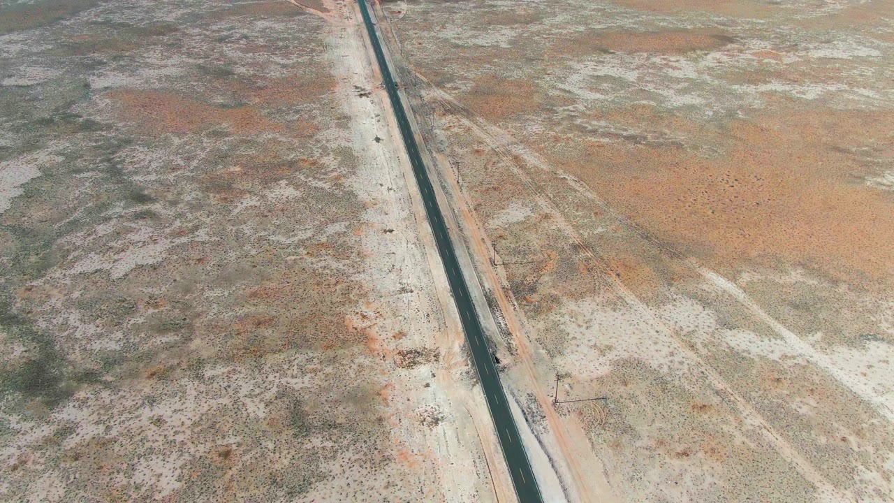 Aerial frame from Guerrero Negro, Baja California Sur, Mexico with the highway nearly vertical in the composition, pale desert on both sides, the truck lower on the road, and no visible people.