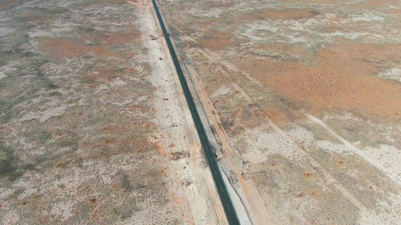 Aerial frame from Guerrero Negro, Baja California Sur, Mexico showing the straight highway shifted toward the right side, mixed pale and orange-brown desert ground, sparse vegetation, and no visible people.