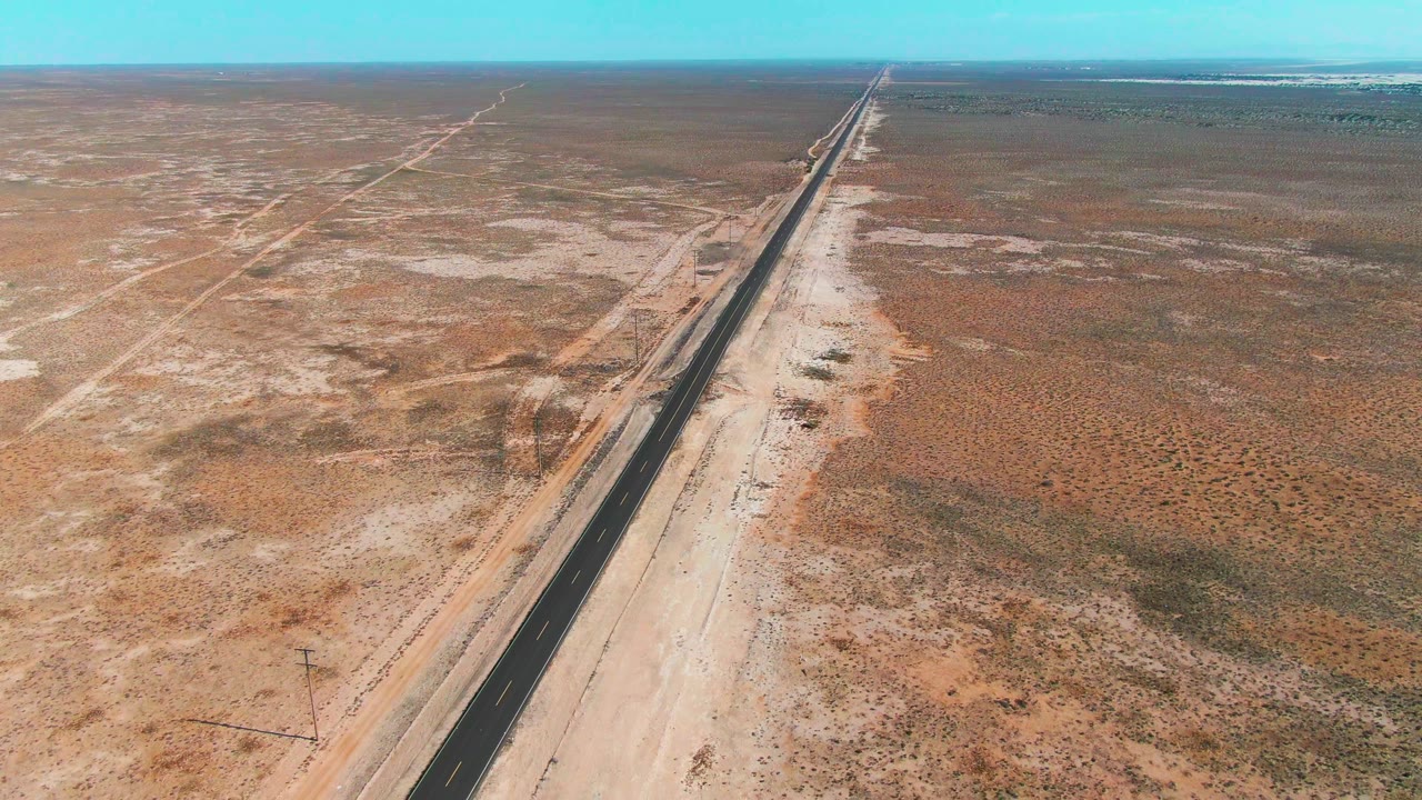 Aerial frame in Guerrero Negro showing the highway slightly farther along the diagonal, bordered by pale sand, scrub patches, and no visible people.
