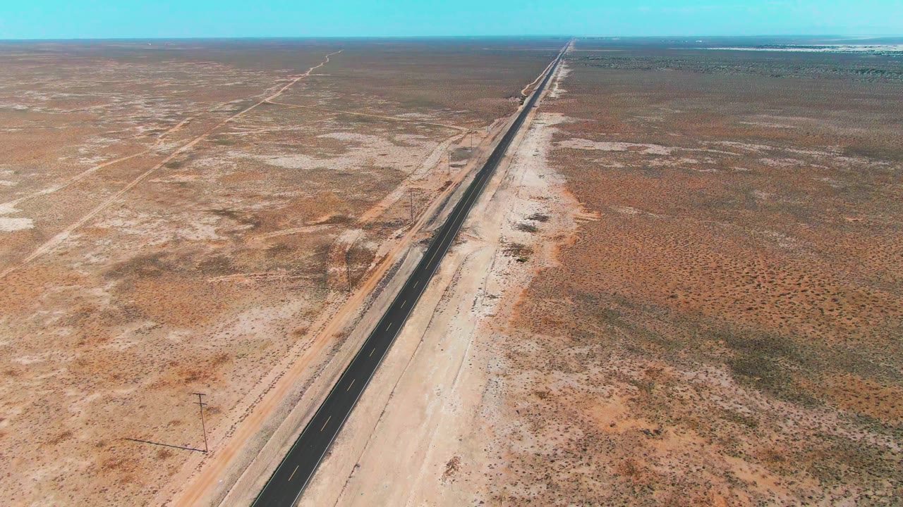 Mid-clip aerial view over Guerrero Negro with the black road, pale shoulders, faint desert tracks, low vegetation, and no visible people.