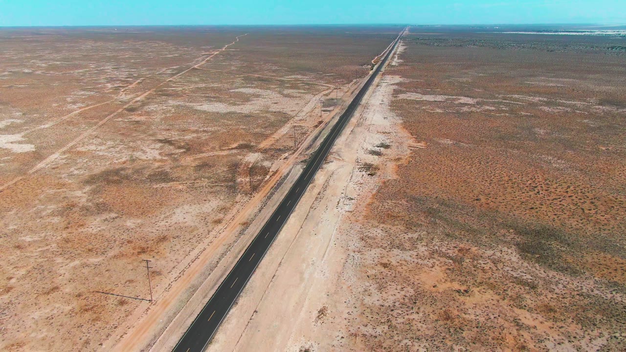 Later aerial frame of the Guerrero Negro desert road with utility poles near the lower edge, sandy ground, scattered scrub, and no visible people.