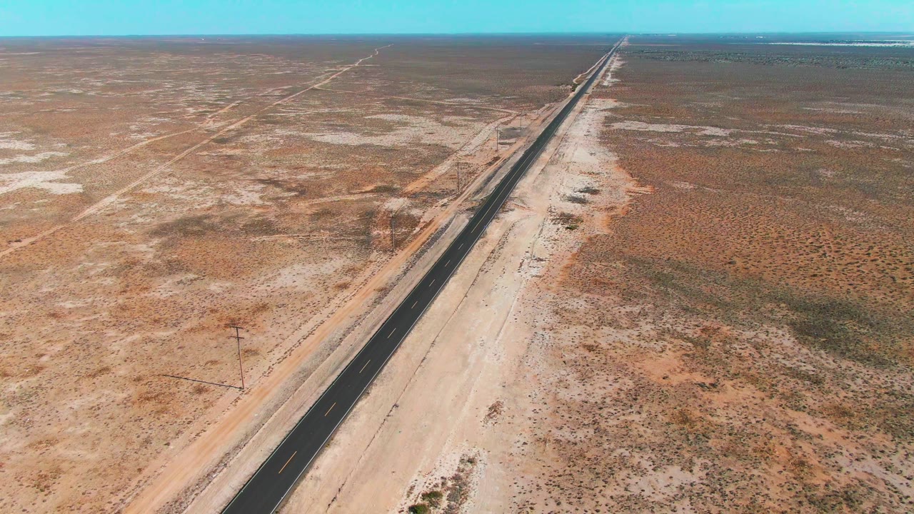 High aerial view in Guerrero Negro, Baja California Sur, with a straight paved highway cutting across sandy desert scrub toward a flat horizon and no visible people.