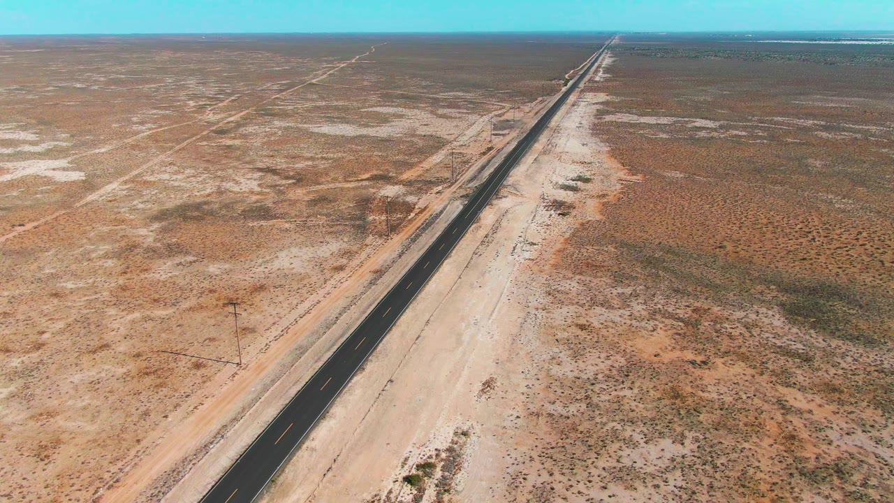 Aerial frame over Guerrero Negro desert terrain, showing the paved highway closer to center with pale shoulders, patchy scrub, utility poles, and no visible people.