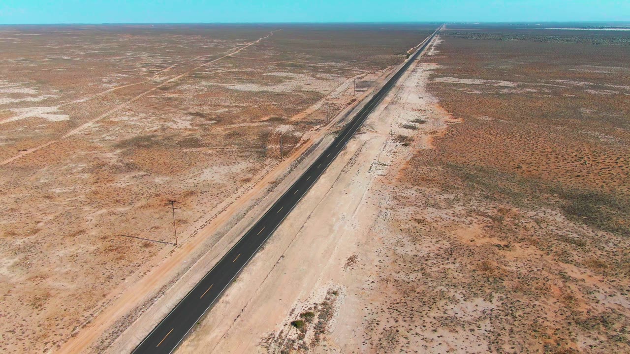 Drone-like aerial view in Guerrero Negro, Baja California Sur, with the road extending from the lower frame toward the far horizon across tan and rust desert ground, no visible people.